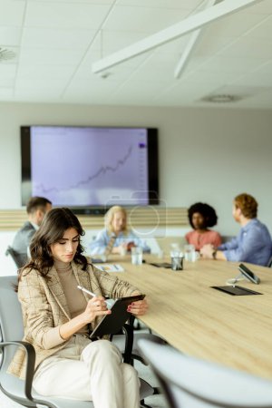 Foto de Empresaria enfocada con un bloc de notas durante una reunión de equipo, con colegas involucrados en la discusión en segundo plano - Imagen libre de derechos