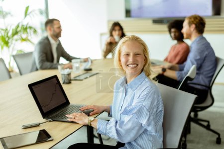 Foto de Alegre mujer de negocios con una computadora portátil sonríe a la cámara mientras sus colegas discuten en el fondo. - Imagen libre de derechos