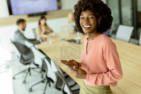 Foto de Ccheerful mujer afroamericana lidera una reunión, agarrando una tableta digital mientras sus colegas participan en la discusión detrás de ella. - Imagen libre de derechos