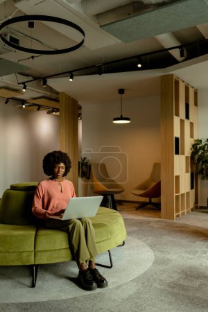 Foto de Mujer de negocios afroamericana animada sonriendo mientras trabaja en su computadora portátil, cómodamente sentada en un vibrante salón de oficina. - Imagen libre de derechos