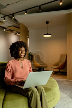 Photo for Spirited African American businesswoman smiling while working on her laptop, comfortably seated in a vibrant office lounge. - Royalty Free Image