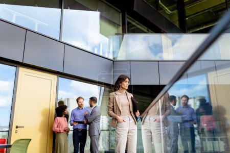 Foto de Una mujer profesional equilibrada camina a través de un espacio de oficina con paredes de vidrio mientras sus colegas conversan en el fondo. - Imagen libre de derechos
