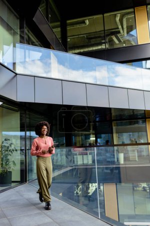 Téléchargez les photos : Femme d'affaires afro-américaine avec un sourire radieux se promène vivement par un immeuble de bureaux, exsudant confiance et élégance dans sa tenue élégante - en image libre de droit