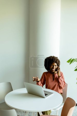 Foto de Una mujer alegre habla en su teléfono en una mesa de café con un ordenador portátil y una planta vibrante a su lado. - Imagen libre de derechos