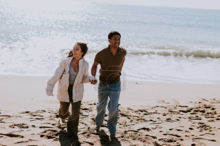 Téléchargez les photos : Un couple souriant profite d'une course insouciante sur un rivage sablonneux, les vagues se brisent doucement en arrière-plan - en image libre de droit