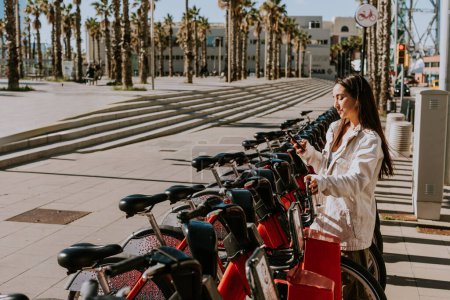 Photo for Young woman stands by a row of red bikes in sunny Barcelona, checking her phone, likely using a bike-sharing app - Royalty Free Image