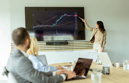 Businesswoman points at growth trends on a screen during a financial meeting, with attendees listening intently