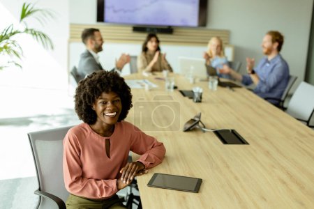 Young female African American professional beams with confidence at a meeting table, colleagues in discussion behind her.