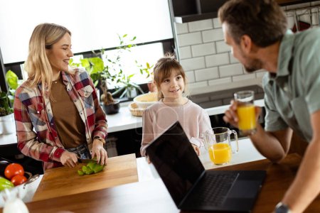Foto de Una familia alegre disfruta de una cálida conversación en su cocina con jugo fresco y un portátil sentado en la mesa. - Imagen libre de derechos
