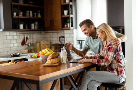 A cheerful couple enjoys a light-hearted moment in their sunny kitchen, working on laptop surrounded by a healthy breakfast