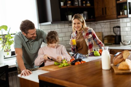 Foto de Una familia prepara una comida juntos, con papá ayudando a su hija a cortar verduras y mamá disfrutando de un jugo. - Imagen libre de derechos