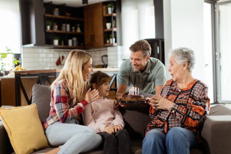 Foto de Escena conmovedora se desarrolla como una familia multigeneracional se reúne en un sofá para presentar un pastel de cumpleaños a una abuela encantada, creando recuerdos para apreciar - Imagen libre de derechos
