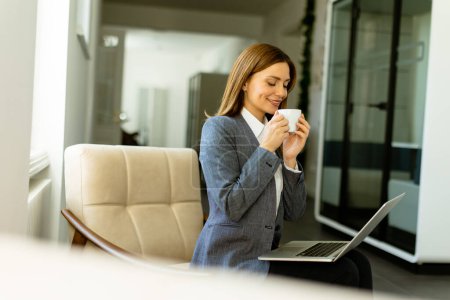 Photo for Poised professional sips her coffee while engaging with her laptop, basking in soft daylight. - Royalty Free Image