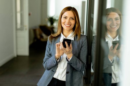 Téléchargez les photos : Une femme professionnelle joyeuse se tient dans un couloir de bureau, son reflet de contenu visible alors qu'elle saisit un smartphone. - en image libre de droit