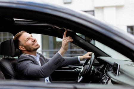 Foto de Hombre sonriente en un traje opera su mecanismo de techo convertibles mientras está estacionado en una carretera urbana - Imagen libre de derechos