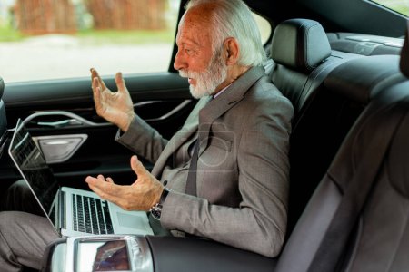 Photo for Elderly executive reacts with astonishment while engaging in a virtual meeting from the backseat of a luxury vehicle. - Royalty Free Image