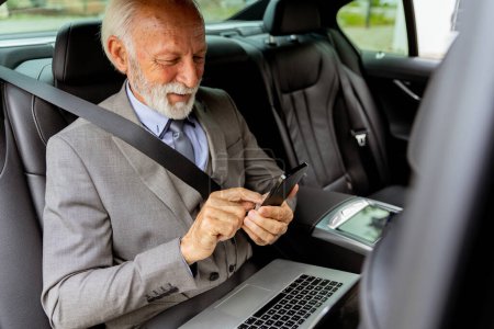 Foto de Distinguido caballero mayor en un traje revisa los mensajes en su móvil durante un viaje en coche, su computadora portátil cerca - Imagen libre de derechos