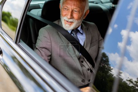 An elderly man with a beard wearing a suit and tie, secured with a seatbelt, looking content inside his vehicle