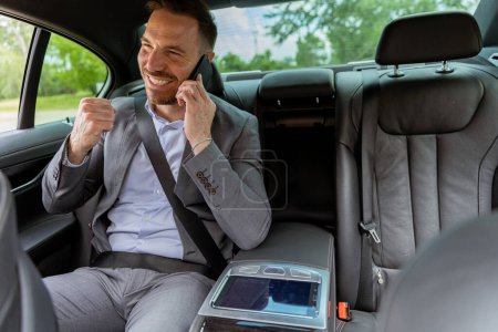 Téléchargez les photos : Homme souriant dans un costume engagé dans un appel animé alors qu'il était assis sur le siège arrière moelleux d'un véhicule de luxe - en image libre de droit
