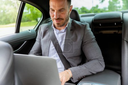 Foto de Hombre enfocado en ropa de negocios escribiendo en un portátil en el asiento trasero de un coche, aprovechando cada minuto - Imagen libre de derechos
