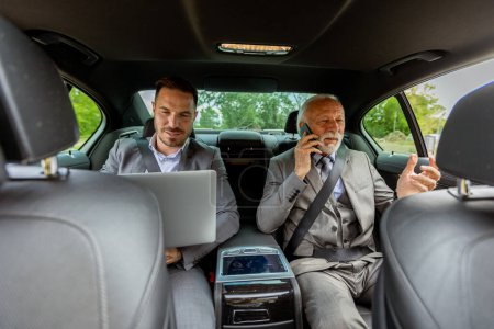 Téléchargez les photos : Deux hommes d'affaires en tenue formelle conversent sur le siège arrière d'une voiture, l'un engagé dans un appel téléphonique - en image libre de droit