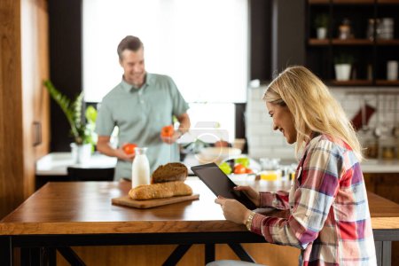 Photo for Woman reads from a tablet at the kitchen counter while a man holding an tomatoes smiles at her - Royalty Free Image