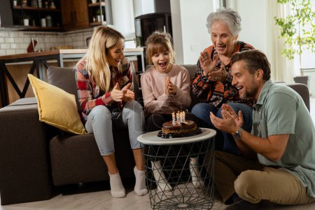 Heartwarming scene unfolds as a multi-generational family gathers on a couch to present a birthday cake to a delighted grandmother, creating memories to cherish