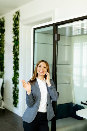 Femme d'affaires bavarde sur son téléphone, son sourire suggérant une conversation agréable au milieu d'un lieu de travail lumineux et élégant