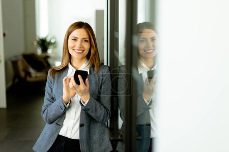 Mujer de negocios chats en su teléfono, su sonrisa que sugiere una conversación agradable en medio de un lugar de trabajo brillante y elegante