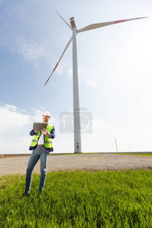 Foto de Un ingeniero de casco de seguridad con portapapeles examina el funcionamiento de una majestuosa turbina eólica en medio de una exuberante vegetación bajo un cielo despejado. - Imagen libre de derechos