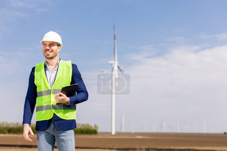 Photo for A cheerful engineer in safety gear reviews data on a tablet before towering windmills under a clear sky. - Royalty Free Image