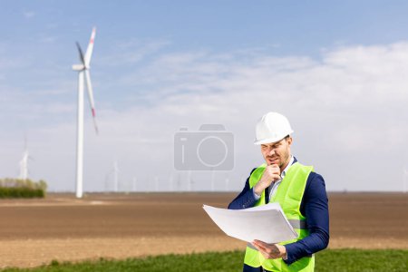 Photo for A cheerful man in safety gear scrutinizes blueprints with windmills towering in the background. - Royalty Free Image