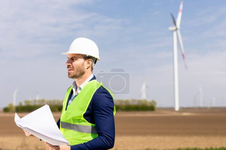 Foto de Un hombre alegre con equipo de seguridad examina los planos con molinos de viento en el fondo. - Imagen libre de derechos