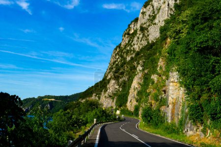 Escénica carretera sinuosa abraza los majestuosos acantilados de Djerdap, Serbia, bajo un cielo azul brillante, con una exuberante vegetación que contrasta con las escarpadas formaciones rocosas
