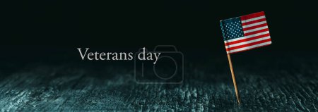 Photo for An American flag, attached to a wooden pole, standing on a dark wooden surface and the text Veterans day against a black background, in a panoramic format to use as web banner or header - Royalty Free Image
