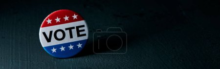 una insignia de voto para las elecciones de Estados Unidos en una superficie gris oscura, con un poco de espacio en blanco a la derecha, en un formato panorámico para su uso como banner web o encabezado