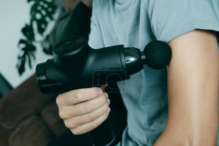 Téléchargez les photos : Un jeune homme caucasien à la maison utilise un pistolet de massage pour masser les muscles de son bras, assis sur le canapé de son salon - en image libre de droit