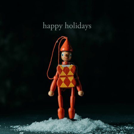 Téléchargez les photos : Le texte joyeuses fêtes et un ornement d'arbre de Noël en forme d'une marionnette à fils arlequin sur un tas de neige, sur un fond noir - en image libre de droit