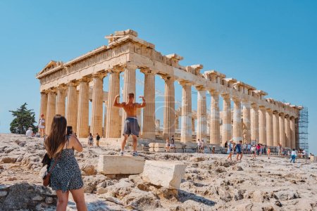 Photo for Athens, Greece - August 30, 2022: A visitor man poses for a photo showing muscle in front of the remains of the famous Parthenon, in the Acropolis of Athens, Greece - Royalty Free Image