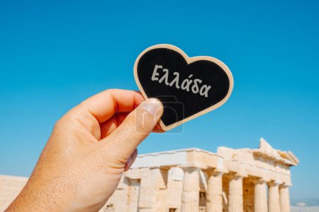 Téléchargez les photos : Gros plan d'un homme tenant un signe en forme de cœur avec le mot Grèce écrit en grec devant le Parthénon, dans l'Acropole d'Athènes, par une journée ensoleillée - en image libre de droit