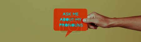 Photo for Holds a paper sign in the shape of a speech balloon with the text ask me about my pronouns on a green background, in a panoramic format to use as web banner - Royalty Free Image