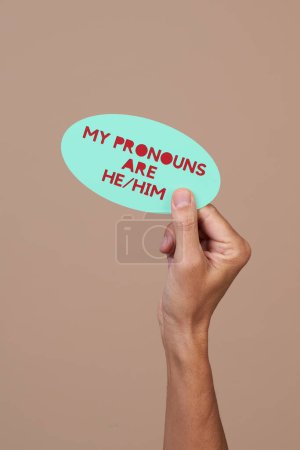 Photo for A person holds a green sign with the text my pronouns are he, him against a brown background with some blank space on top - Royalty Free Image