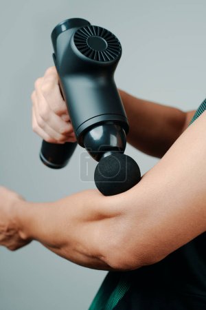 Foto de Primer plano de un joven, en ropa deportiva, usando una pistola de masaje para masajear los músculos de su brazo, cerca de su codo - Imagen libre de derechos