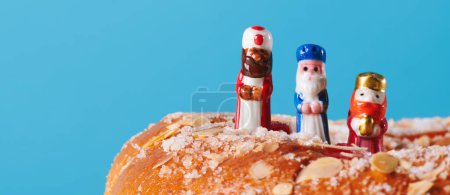 the three wise men, melchior, caspar and balthazar, on top of a roscon de reyes, the spanish king cake, on a blue background in a panoramic format to use as web banner