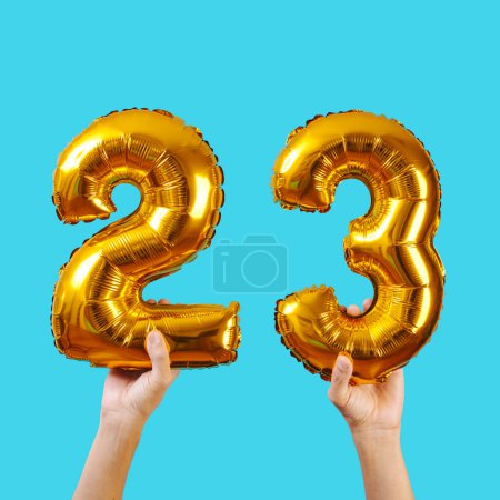 Foto de Hombre sostiene dos globos dorados en forma de número que forman el número 23 sobre un fondo azul - Imagen libre de derechos