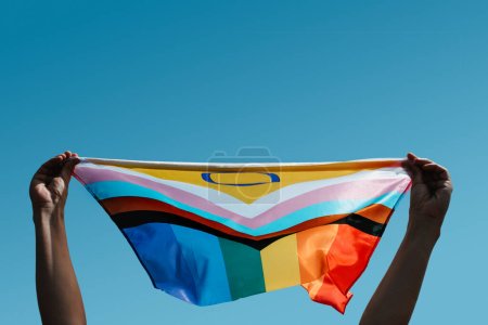 closeup of a young person holding an intersex-inclusive progress pride flag above their head against the blue sky, with some blank space on top