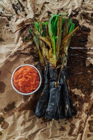 Photo for High angle view of some roasted calcots, sweet onions typical of Catalonia, Spain, and a white ceramic bowl with some romesco sauce to dip them in it, on a crumpled kraft paper - Royalty Free Image