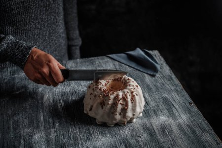Foto de Un hombre caucásico corta un delicioso pastel de gugelhupf, cubierto con un glaseado de vainilla, con un cuchillo de cocina, en una mesa de madera rústica gris - Imagen libre de derechos