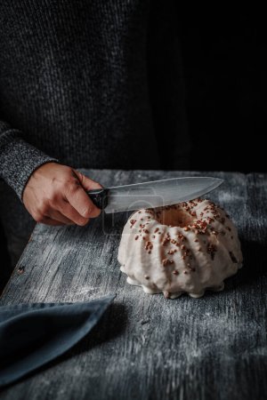 Photo for Closeup of a man cutting a delicious gugelhupf cake covered with a vanilla frosting, placed on a gray rustic wooden table - Royalty Free Image