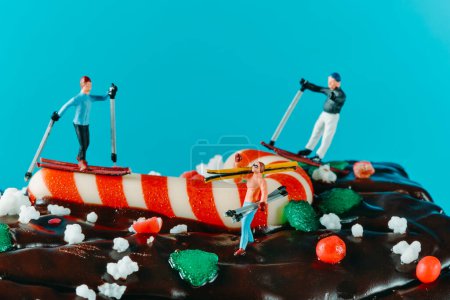 Téléchargez les photos : Gros plan de quelques personnes miniatures skiant ou portant leurs skis sur un gâteau en rondins de Noël, sur un fond bleu - en image libre de droit